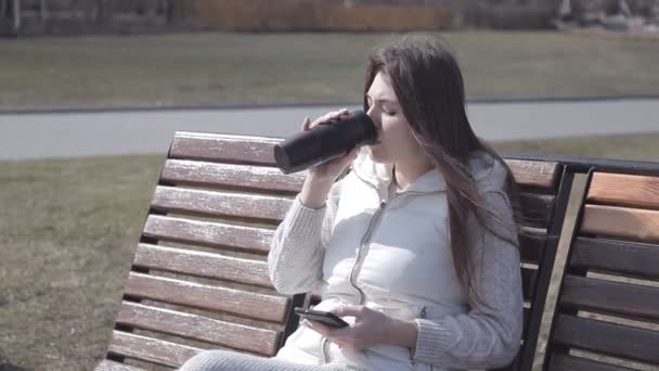 Krásná mladá dívka sedící na dřevěnou lavici v city parku v bílém obleku pití kávy z tepelné hrnek a zadáte do telefonu — Stock video