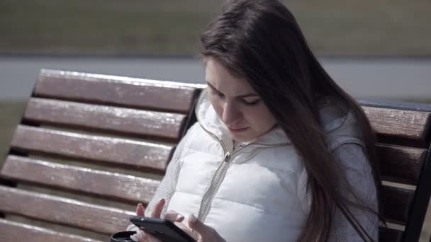 Bela jovem sentada em um banco de madeira em um parque da cidade em um terno branco beber café de uma caneca térmica e digitar no telefone. de perto — Vídeo de Stock