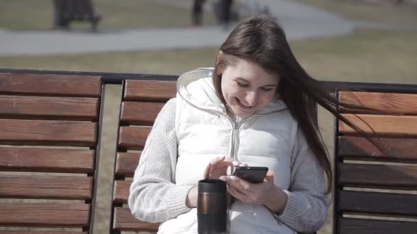 Hermosa joven sentada en un banco de madera en un parque de la ciudad en un traje blanco beber café de una taza térmica y escribir en el teléfono — Vídeo de stock