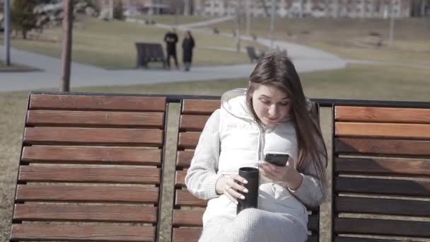 Красивая молодая девушка сидит на деревянной скамейке в городском парке в белом костюме, пьет кофе из тепловой кружки и печатает в телефоне — стоковое видео