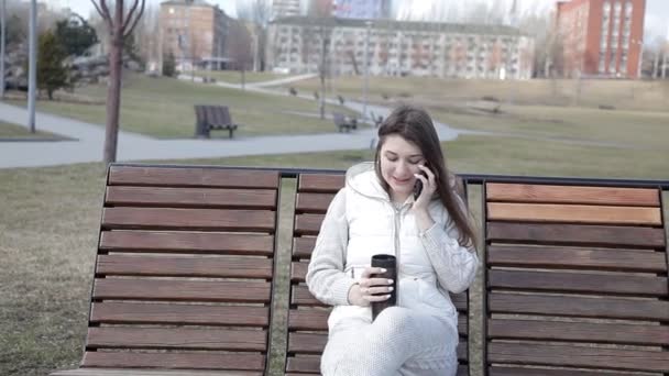 Красивая девушка в парке на скамейке пьет кофе из кружки и разговаривает по телефону — стоковое видео