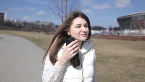 Uma linda garota caminha no parque bebendo café de uma caneca térmica e mensagens de texto no telefone, sorrindo — Vídeo de Stock