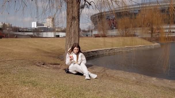 Menina bonita comendo uma maçã vermelha no parque sob uma árvore perto da água e lendo um livro — Vídeo de Stock