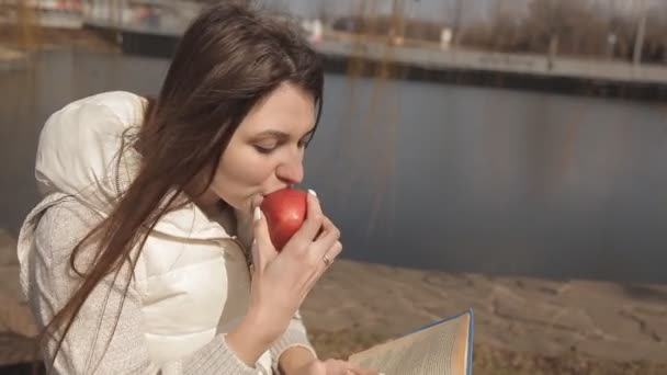 Красивая молодая девушка ест красное яблоко в парке под деревом у воды и читает книгу — стоковое видео