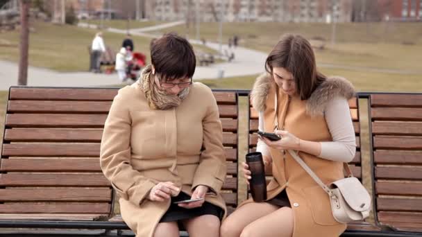 Мать и дочь, сидящие в парке, общаются и используют мобильный телефон. Природа, отношения — стоковое видео