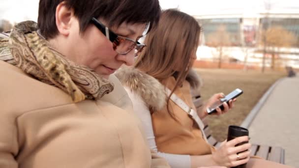 Madre e hija se sientan en el parque en un banco y usan teléfonos móviles — Vídeo de stock