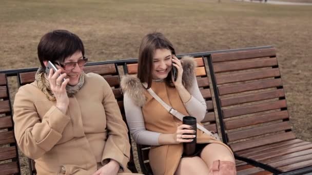 Μητέρα και κόρη κάθονται στο πάρκο σε ένα παγκάκι και μιλούν σε ένα κινητό τηλέφωνο. Οικογένεια, ώρα, περίπατος — Αρχείο Βίντεο