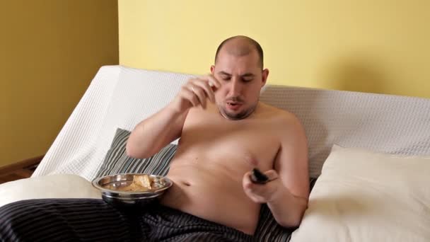 アウターのない怠惰な、ハゲ、ぽっちゃり太った男は、テレビを見て、チップがソファーの上に横たわってコカコーラを飲んでいます。太った男は間違ったライフスタイルを導く — ストック動画
