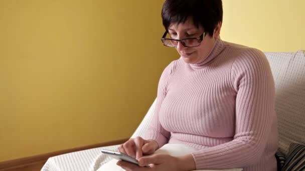 Een vrouw in een bril het schrijven van een bericht op haar gloednieuwe smartphone zittend op de Bank met een wit kussen thuis — Stockvideo