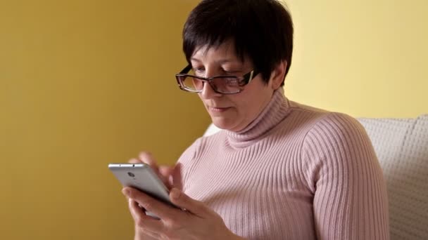 Una mujer con gafas escribiendo un mensaje en su nuevo smartphone mientras está sentada en el sofá con una almohada blanca en casa — Vídeos de Stock