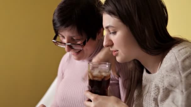 Madre e figlia sedute a casa sul divano. L'uso degli smartphone, la navigazione, la lettura delle notizie. Bevi Coca-Cola, whisky — Video Stock