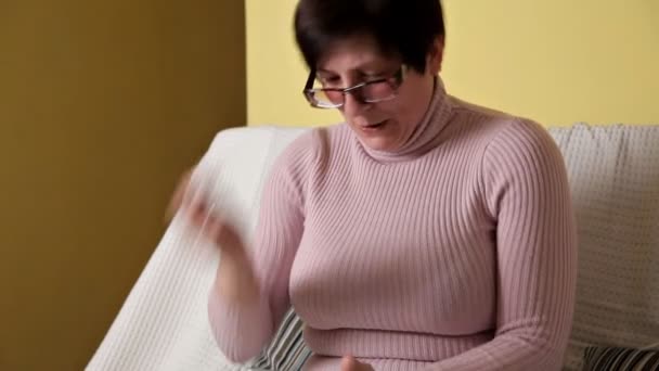 A woman in a pink jacket with glasses sitting on a white couch and texting — Αρχείο Βίντεο