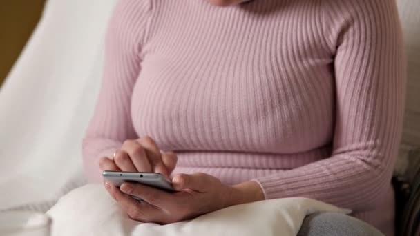 A woman in a pink jacket with glasses sitting on a white couch and texting — Αρχείο Βίντεο