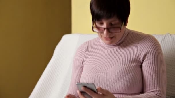 A woman in a pink jacket with glasses sitting on a white couch and texting — Αρχείο Βίντεο