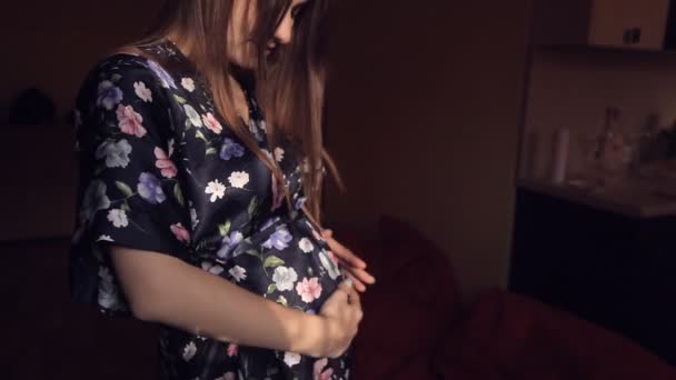 Беременная молодая девушка в шелковом халате осторожно и с любовью гладит живот — стоковое видео