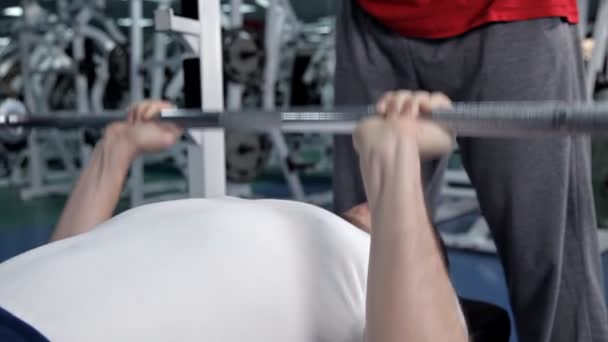 Man raises free weight under control of instructor in sports club. Handsome guy lies on bench and does exercise with iron object — Stock Video