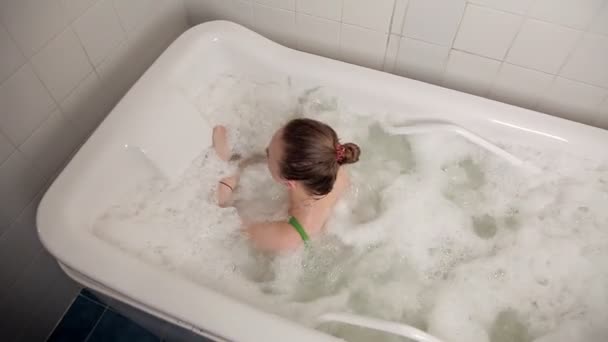 Ein junges Mädchen im Badeanzug verbringt die Zeit im Wellnessbereich, im Badezimmer mit Hydromassage und Schaumstoff liegend. Behandlung, Schönheit, Pflege — Stockvideo