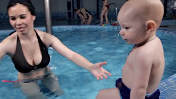 A fiatal anya megtanítja a babát úszni a medencében. Leül a víz mellé, aztán segít neki lemerülni. Aranyos baba. — Stock videók