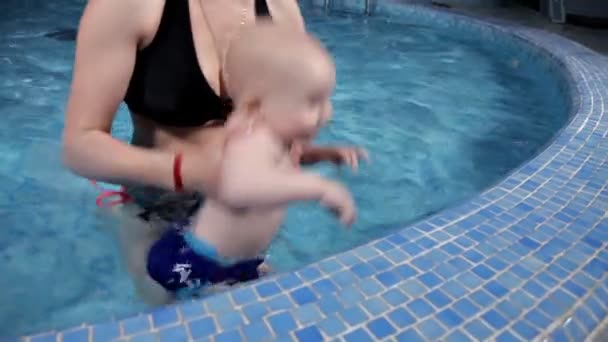 Jonge moeder leert baby zwemmen in het zwembad. Ze zit bij het water en helpt hem duiken. Schattige baby. — Stockvideo