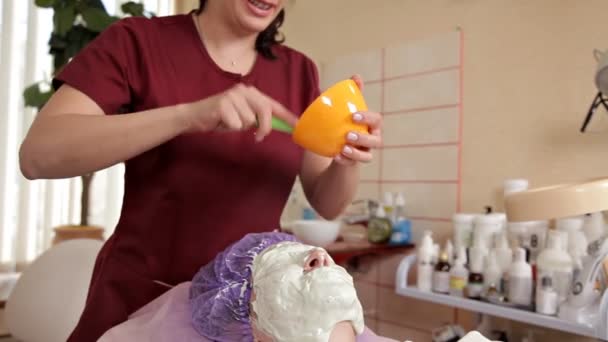 Um cosmetologista experiente prepara uma máscara saudável para o cliente. Ela se levanta e toca a cara com um pincel. Uma jovem deita-se de olhos fechados. Facial pele care.Alginate máscara — Vídeo de Stock