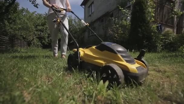 Девушка стрижет неровный газон желтой газонокосилкой босой — стоковое видео