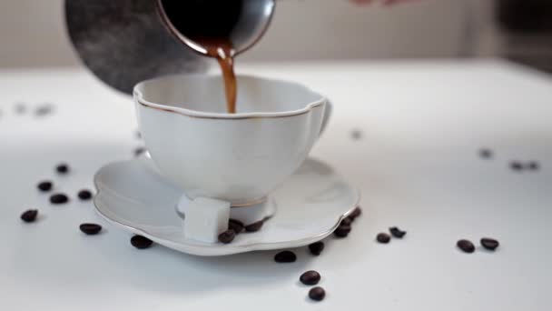 Kvinnans hand häller kaffe från en turkisk kaffe gryta i en vit kopp på ett vitt bord. Kanel och kaffe bönor — Stockvideo