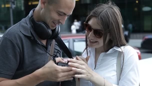 Два мандрівники чоловік і жінка переглядають зняті фотографії на камеру, що стоїть в центрі міста серед хмарочосів — стокове відео