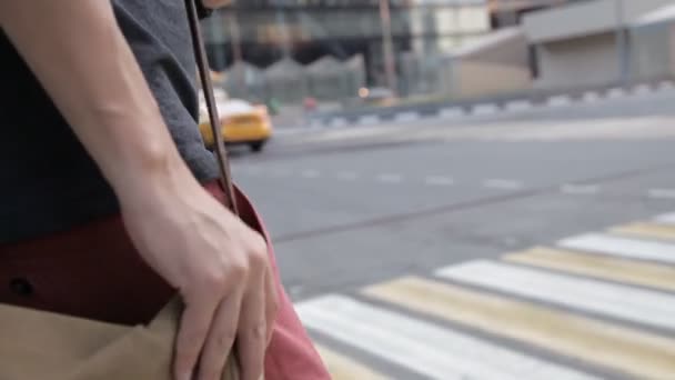 Ein Mann überquert die Straße an einem Fußgängerüberweg, in den Händen einer Kamera auf seiner Umhängetasche. auf dem Hintergrund eines vorbeifahrenden Taxis. Nahaufnahme der Hände — Stockvideo