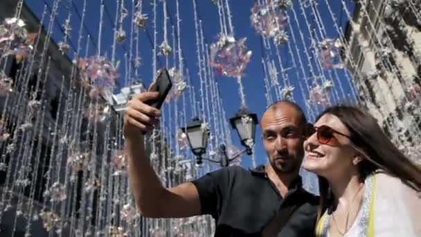 Het meisje en de man maken een foto op de straat op de achtergrond van straatlampen op de draad. Lampen-lampen hangen decoraties tegen de blauwe lucht in de zomer — Stockvideo