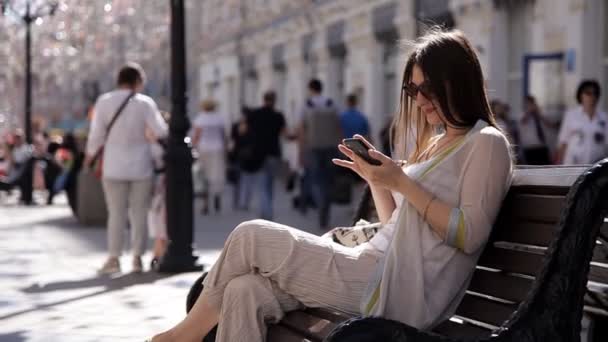 Młoda dziewczyna siedzi na ławce na ulicy na tle pięknej biżuterii i drukuje, pisze wiadomość na smartfonie — Wideo stockowe