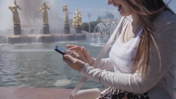 Radosna dziewczyna w beżowym kolorze za pomocą smartfona siedzącego przy fontannie w parku w gorącym lecie — Wideo stockowe