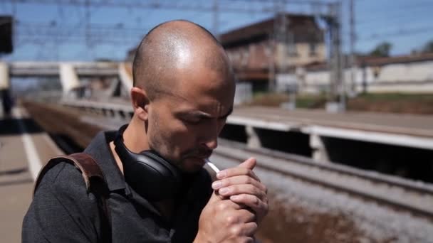 Der charismatische Mann auf dem Bahnsteig des Bahnhofs zündet sich eine Zigarette an und wartet auf den Zug. Mann mit Kopfhörer raucht an einem sonnigen Tag auf dem Bahnsteig. — Stockvideo