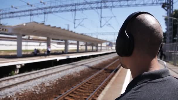 Uomo calvo carismatico che ascolta musica sulle cuffie in attesa di un treno, fumando una sigaretta. Fumare alla stazione ferroviaria — Video Stock