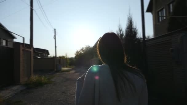 Disparando por la espalda, aguas por la espalda, una chica con el pelo largo está caminando por la calle entre las casas al atardecer — Vídeos de Stock