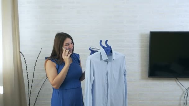 Una joven habla por teléfono y usa vapor para acariciar la camisa del hombre. El proceso de vapor de la ropa al vapor. Detalles azules — Vídeo de stock