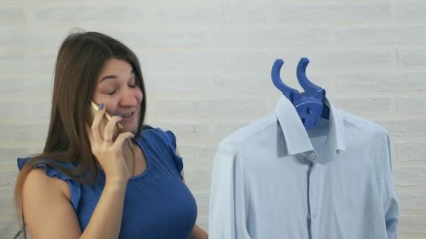 若い女性が電話で話し、男性のシャツをストロークするために蒸気を使用しています。蒸し服の蒸気のプロセス。青の詳細 — ストック動画