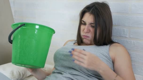 Утренняя тошнота. Молодая беременная женщина лежит на диване и блюет в ведро. Опьянение от беременности — стоковое видео