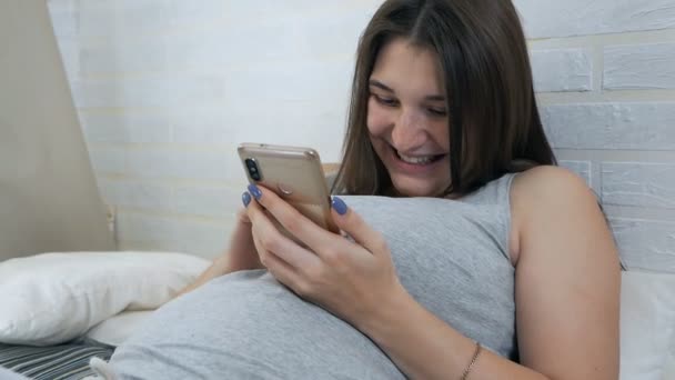 Молодая беременная девушка лежит на диване и пользуется мобильным телефоном. Досуг беременной женщины — стоковое видео