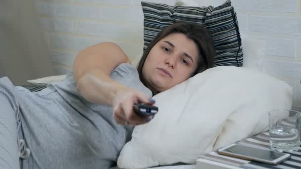 Ragazza incinta assonnata si trova sul divano, guarda la TV e sbadiglia. La donna incinta passa usando il telecomando TV — Video Stock