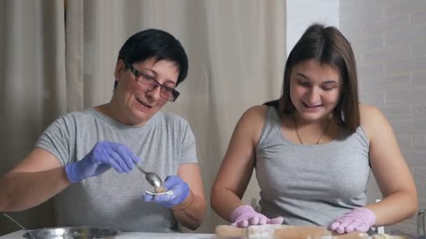 Une fille enceinte et sa mère s'assoient à une table dans la cuisine de la maison et préparent des boulettes avec de la viande hachée pour le déjeuner, déroulent la pâte avec un rouleau à pâtisserie — Video