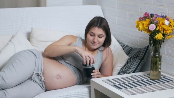 Lycklig gravid kvinna ligger på soffan hemma gäller grädde på magen — Stockvideo