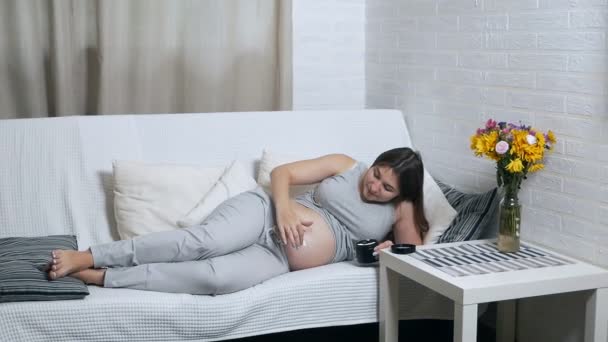 Mutlu hamile bir kadın evdeki kanepede uzanmış, karnına krem sürerek gülümsüyor. — Stok video