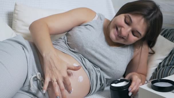 Boldog terhes nő fekszik a kanapén otthon alkalmazza krém a gyomra formájában egy mosolygós — Stock videók