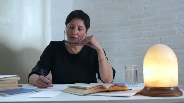 Een vrouwelijke dichter in zwarte jurken en glazen zit aan een tafel en schrijft een gedicht, gooit een bruisende tablet en vitaminen voor hersenactiviteit in een glas water. Oprichting — Stockvideo