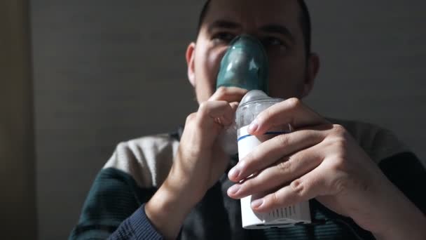 Jonge man met een masker uit een inhaleertoestel thuis. Ontsteking van de luchtwegen via vernevelaar behandelt. Preventie van astma en hoesten — Stockvideo