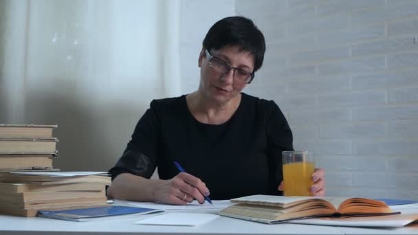 Une femme en robe noire s'assoit à une table sur fond blanc et prend des notes sur papier, boit un verre d'eau et des vitamines pour l'activité cérébrale. Poétesse, poète, créativité, livres — Video