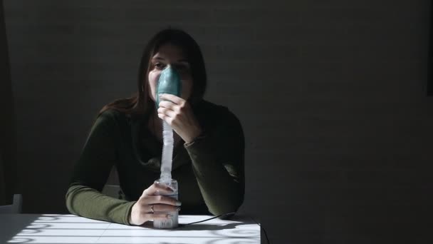Используйте для лечения ингалятор и распылитель. Молодая женщина сидит за столом, вдыхая ингалятор через маску на темном фоне — стоковое видео