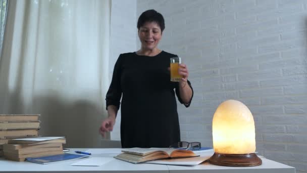 En kvinna i svart klänning sitter och sitter vid ett bord på en vit bakgrund och antecknar på papper, dricker ett glas vatten och vitaminer för hjärnaktivitet. Poesi, poet, kreativitet.books — Stockvideo
