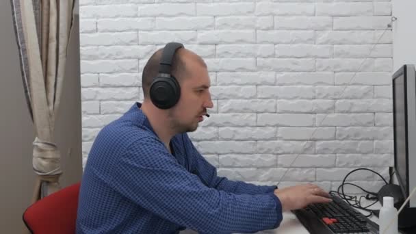 Молодой фрилансер, работающий из дома за компьютером. Человек на удаленной работе сидит в халате и наушниках за компьютером на самоизоляции в случае вируса — стоковое видео