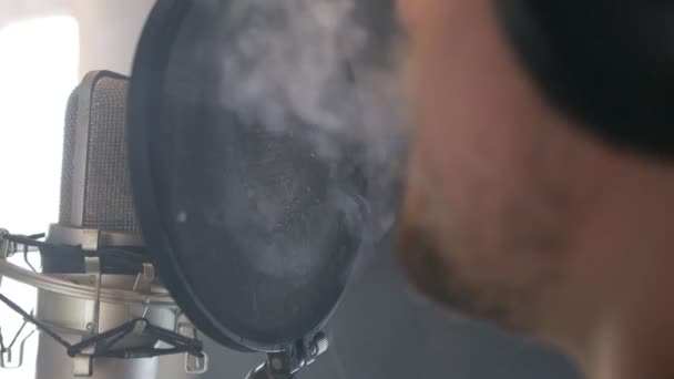 Рэп-артист в наушниках поёт песню в микрофон в студии звукозаписи, курит и пускает дым. Шоу-бизнес. Рецептурная студия — стоковое видео